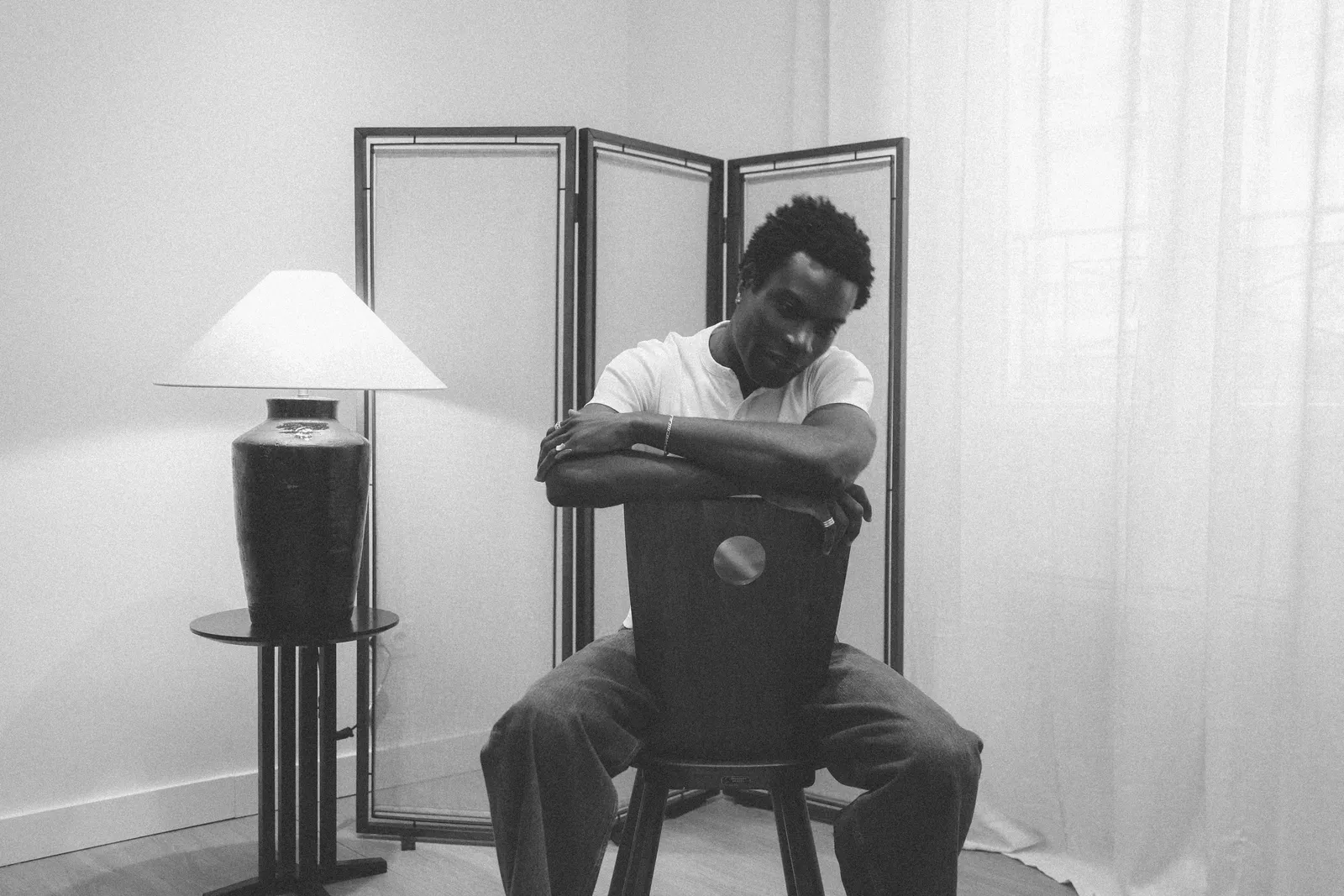 Photographie en noir et blanc : une personne assise sur une chaise, près d'une lampe et d'un paravent.